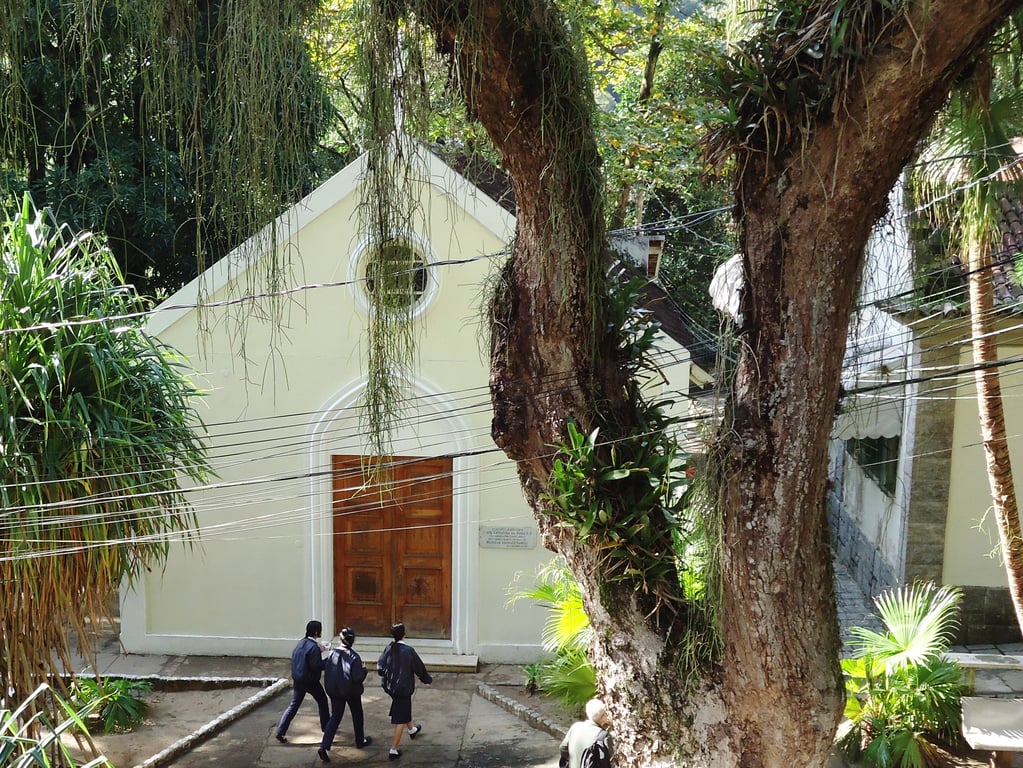 Fachada de escola com características militares, com bandeira do Brasil e vegetação na frente, em dia ensolarado