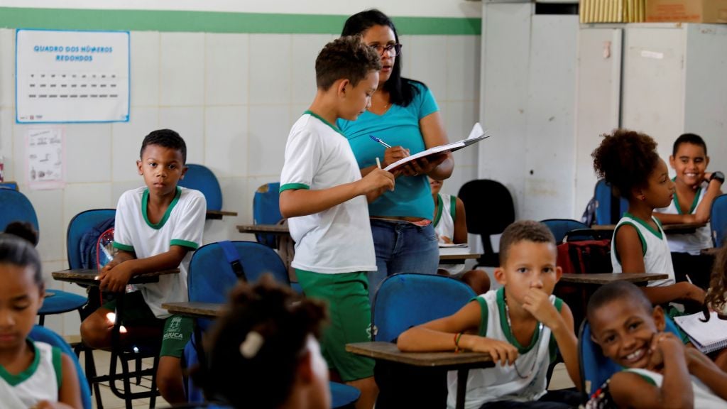 Sala de aula em escola pública brasileira, com professor e estudantes em atividade, luz natural e quadro verde