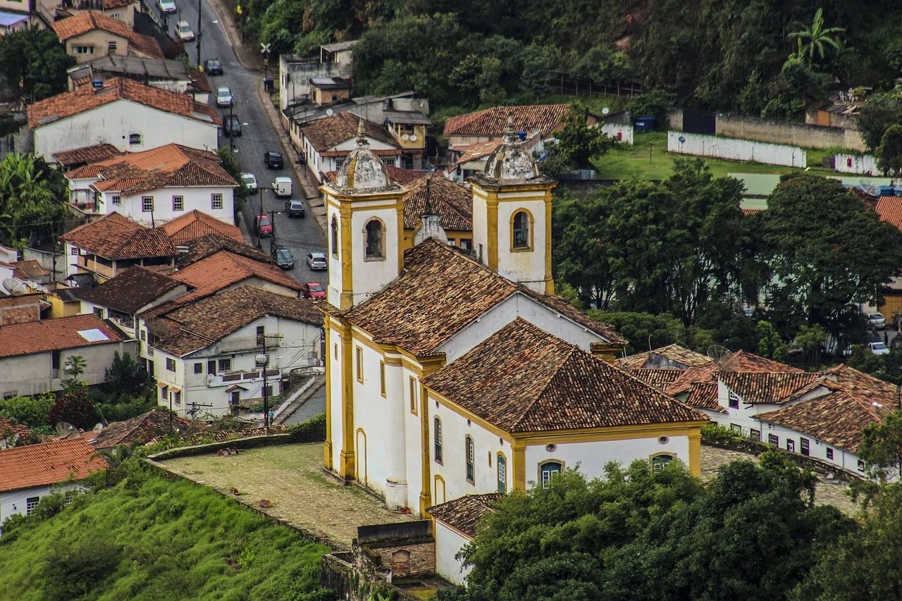 Vista aérea de cidade histórica em Minas Gerais com igreja e casario, serras ao fundo, céu aberto e luz de fim de tarde