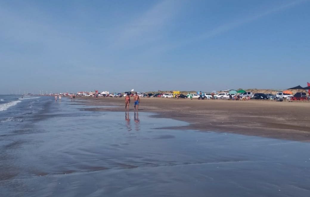 Praia do Cassino, com longa faixa de areia e mar; pessoas caminhando e carros estacionados ao fundo, céu azul.