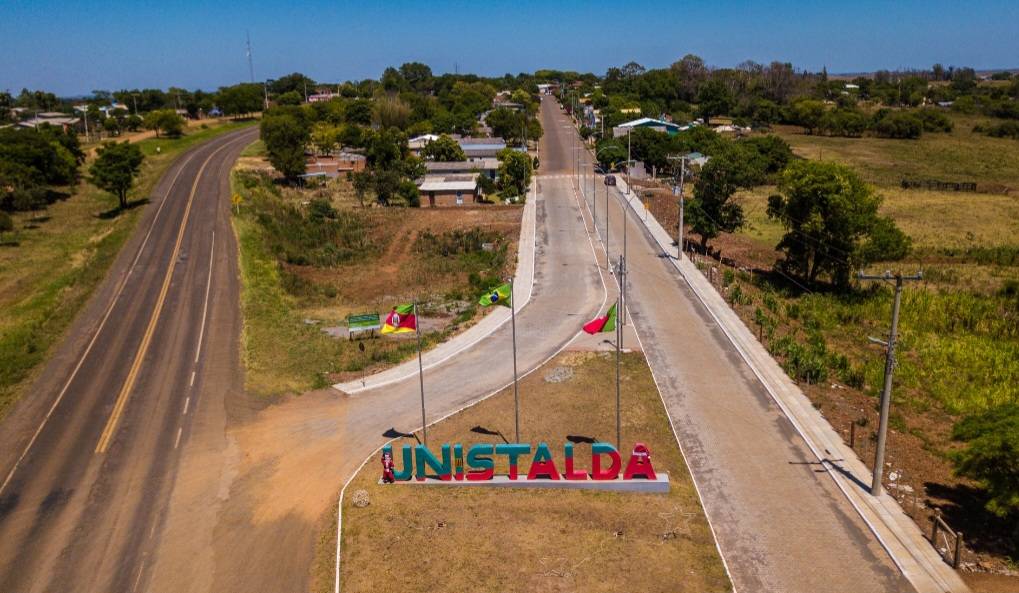 Entrada da cidade com totem “UNISTALDA” e bandeiras ao fundo, em via tranquila de zona rural