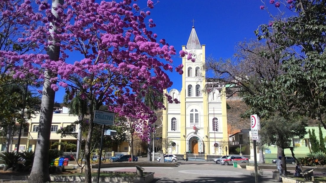 Vista da praça central de Cláudio MG em dia ensolarado, com igreja ao fundo e árvores floridas