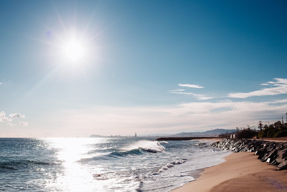 Orla de praia ao entardecer, com mar azul, faixa de areia e vegetação de restinga, em composição horizontal