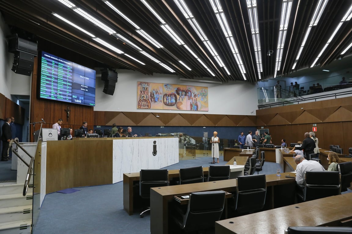 Interior de uma câmara legislativa brasileira, com bancadas, microfones, painel e bandeiras