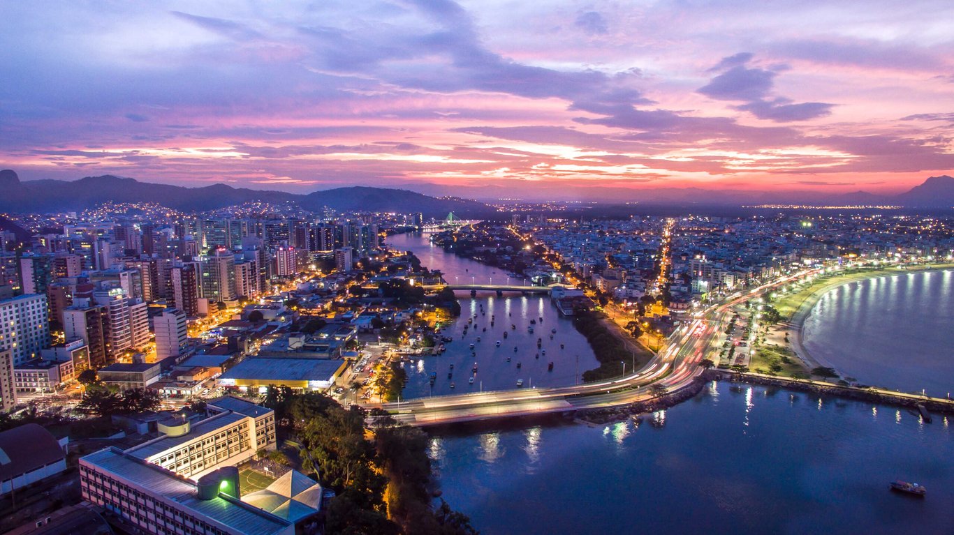 Vista aérea de Vitória ES ao pôr do sol, com a Terceira Ponte e a baía ao fundo.