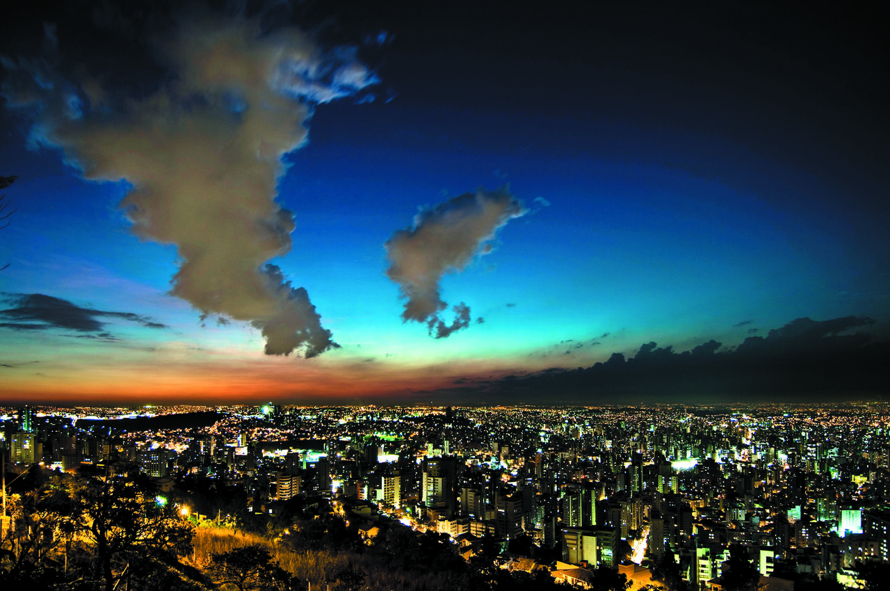 Vista aérea de Belo Horizonte ao entardecer, com edifícios iluminados e montanhas ao fundo.