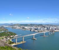 Vista aérea de Vitória ES, com a Baía de Vitória e a Terceira Ponte ao fundo, em dia claro.