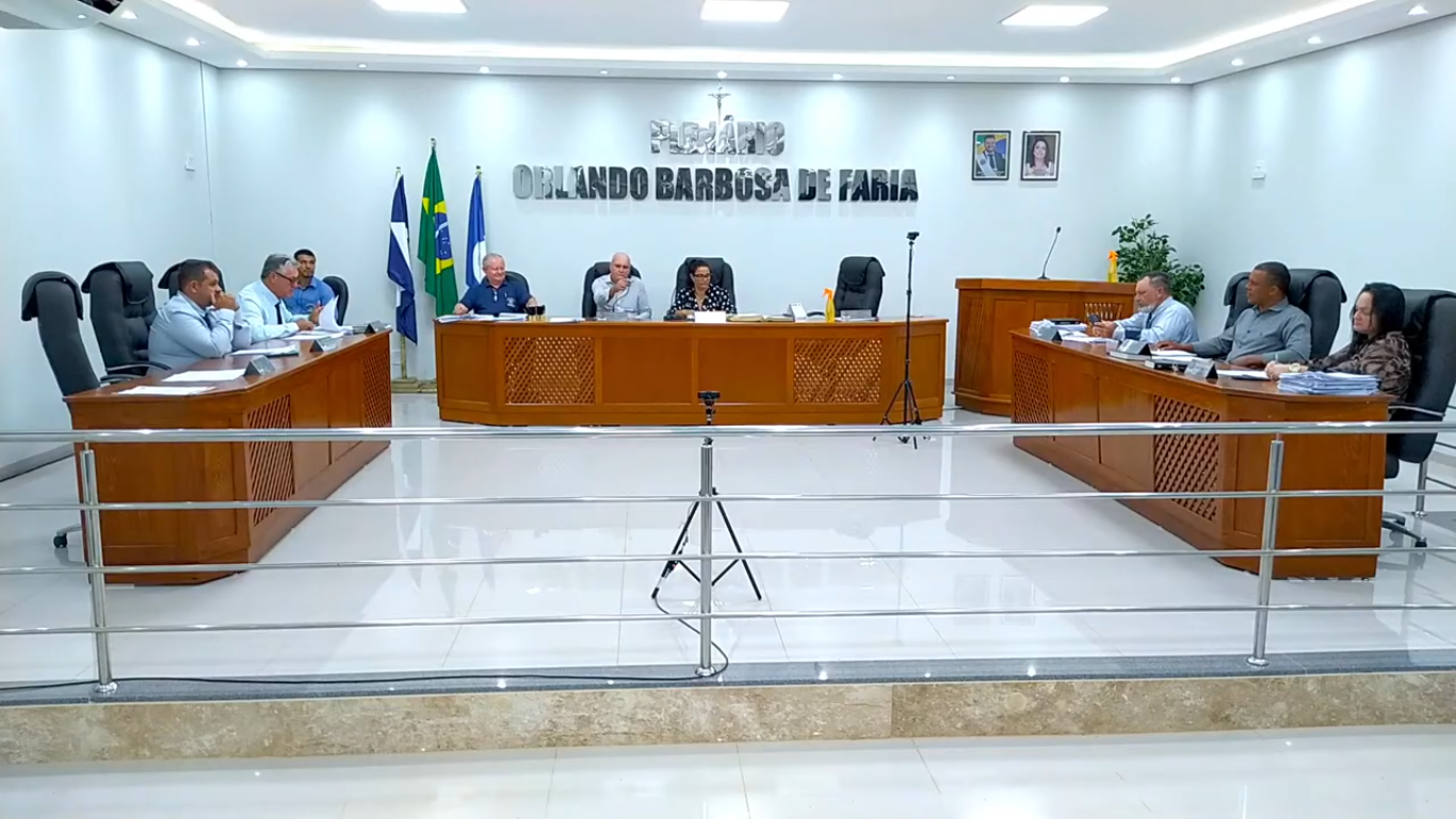 Plenário de câmara municipal brasileira, com cadeiras, mesa diretora e brasão ao fundo