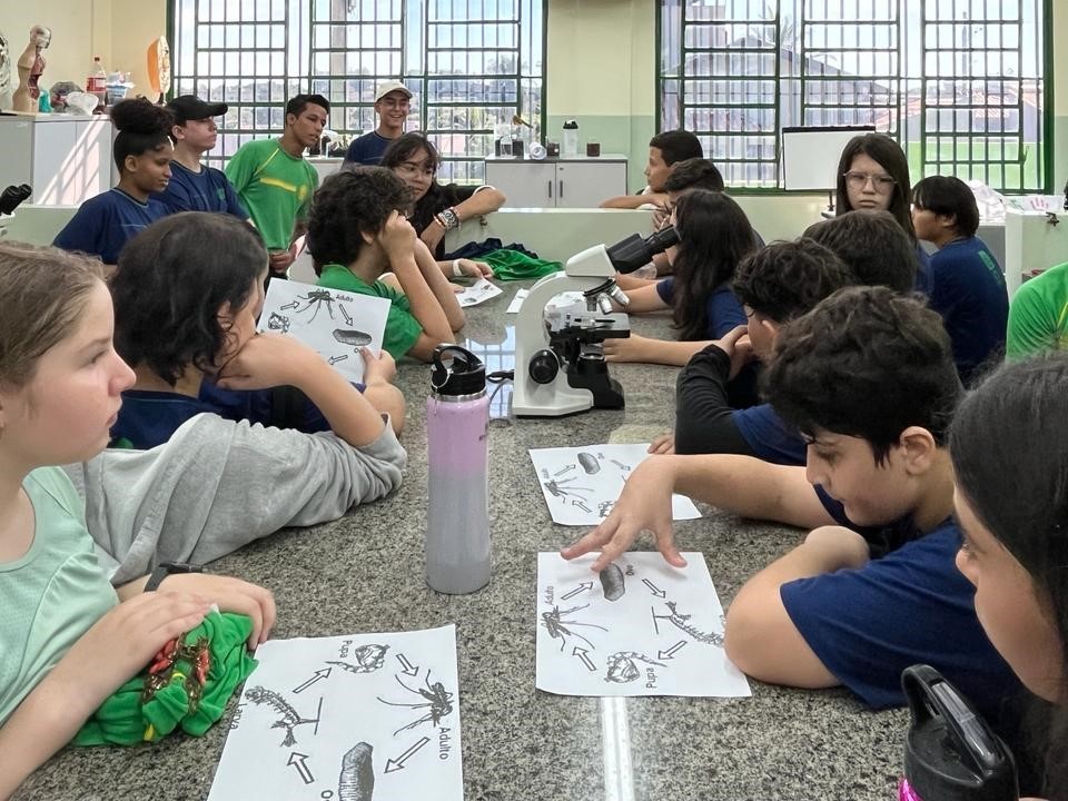 Foto de um laboratório de química educacional com alunos e professor. Os estudantes observam materiais didáticos em uma bancada.