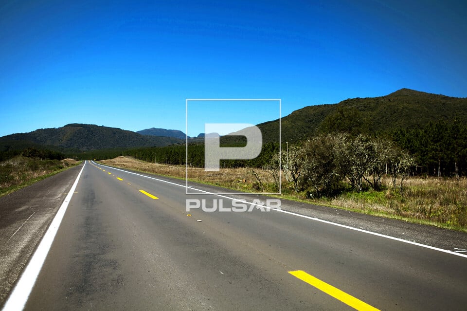Estrada na região serrana de Santa Catarina, vegetação densa e morros ao fundo sob céu azul