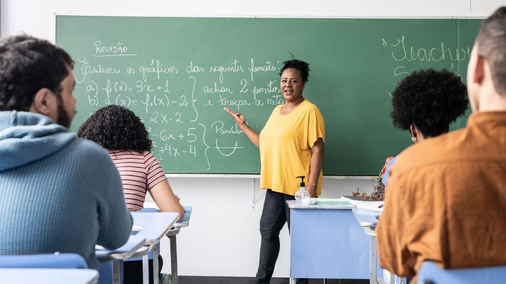 Sala de aula do ensino fundamental com professora em atividade, alunos sentados e quadro ao fundo