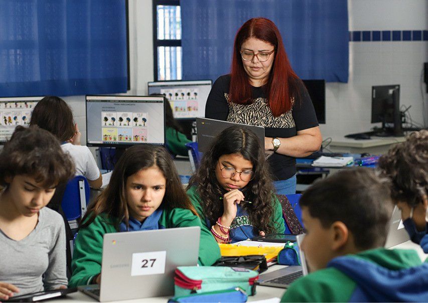 Sala de aula moderna com alunos em computadores e professor supervisionando