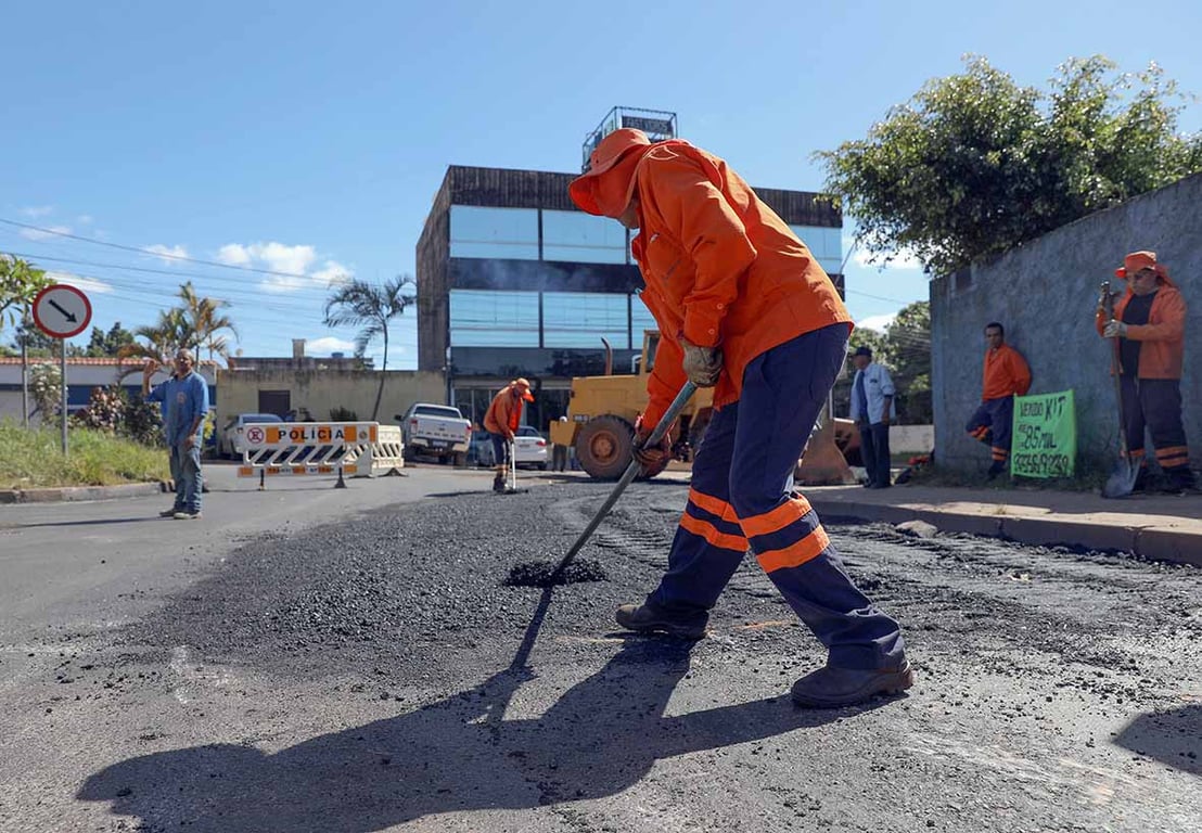 Pavimentação asfáltica em operação, com rolo compactador e usina ao fundo, em cenário urbano-rural