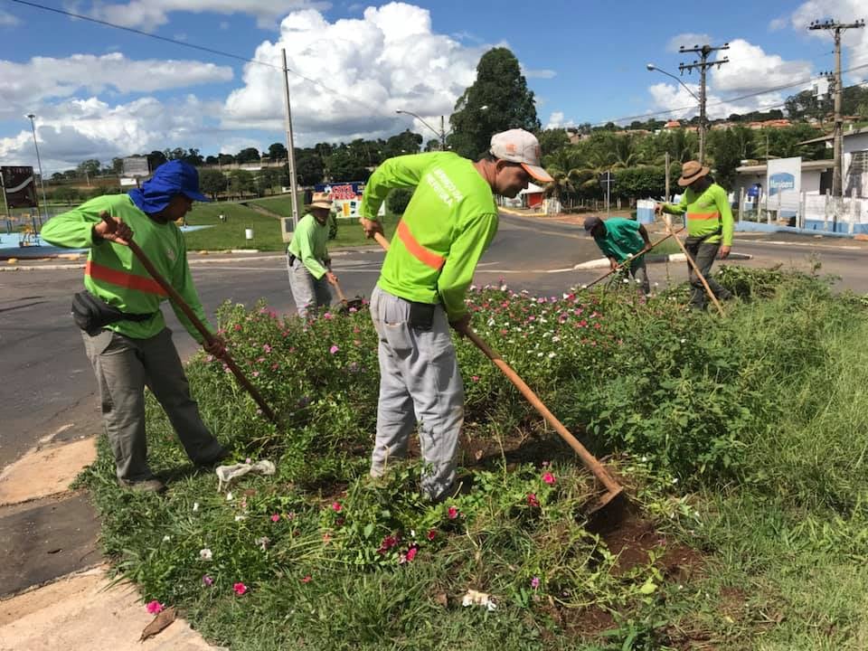 Equipe de jardinagem municipal realizando manutenção de canteiro de flores, com ferramentas de poda e roçadeira