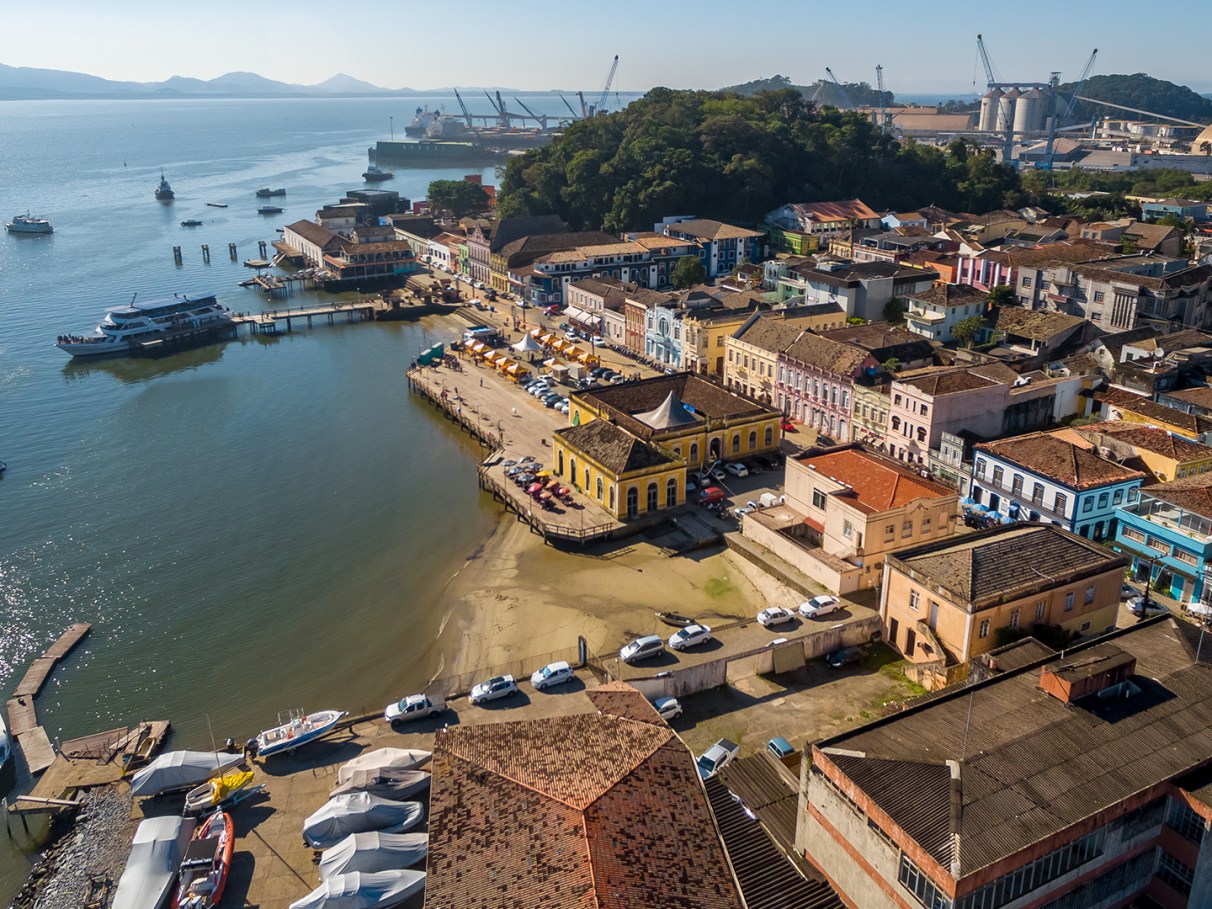 Vista aérea do centro histórico de São Francisco do Sul, evidenciando a arquitetura colonial à beira da baía. A cena apresenta barcos e construções coloridas.
