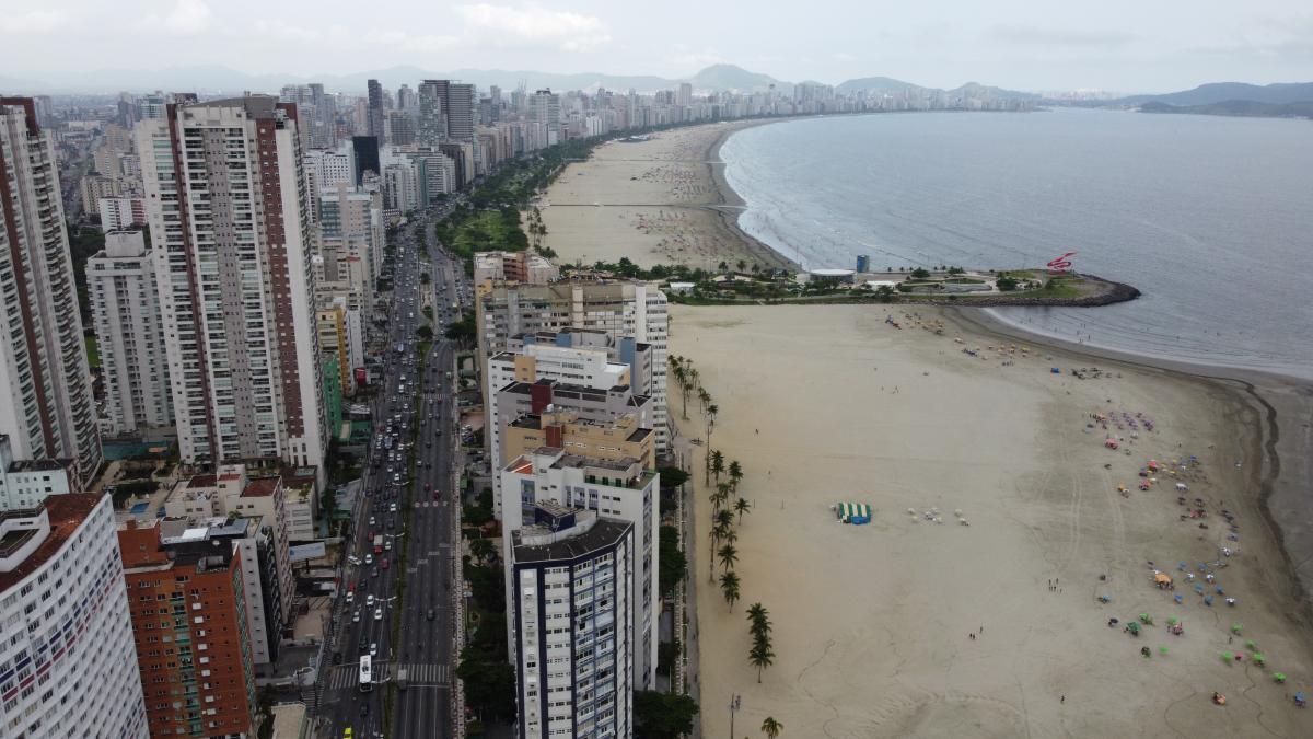 Vista aérea da orla de Santos com jardins e prédios residenciais; praia e mar sob céu aberto.