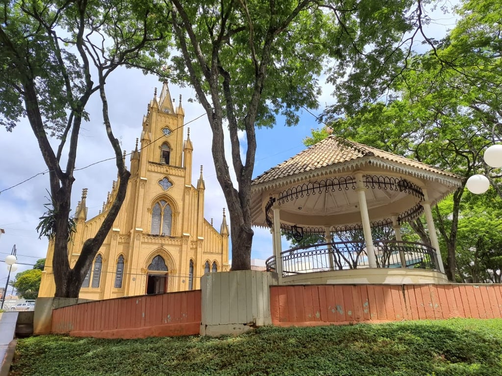 Praça central de Carmo da Mata com igreja histórica e coreto ao fundo, em dia parcialmente nublado