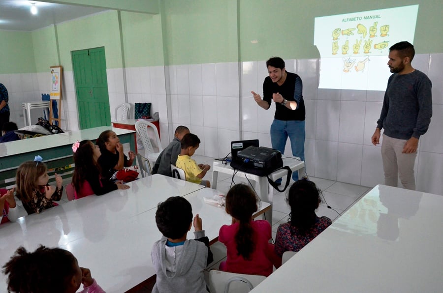 Sala de aula com intérprete de Libras ao lado do professor; estudantes atentos acompanham a explicação