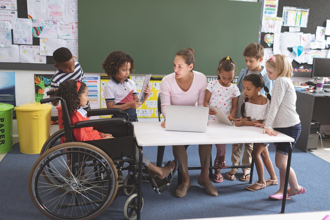 Foto de uma sala de aula com uma professora e alunos envolvidos em atividades. Crianças se reúnem ao redor de um computador e mostram trabalhos.