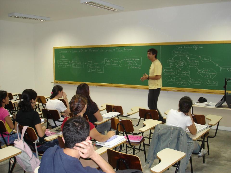 Sala de aula universitária com alunos e professor em atividade; quadro ao fundo com anotações