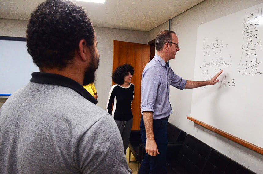 Professor em sala de aula universitária, escrevendo no quadro enquanto alunos acompanham