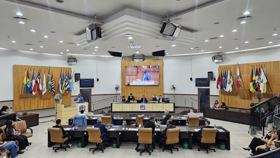 Interior de uma câmara municipal com mesas, cadeiras e bandeiras; sessão legislativa em andamento