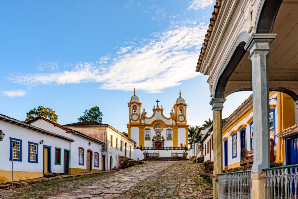 Vista urbana com igreja e casario histórico, céu azul, representando cidade do interior de Minas Gerais