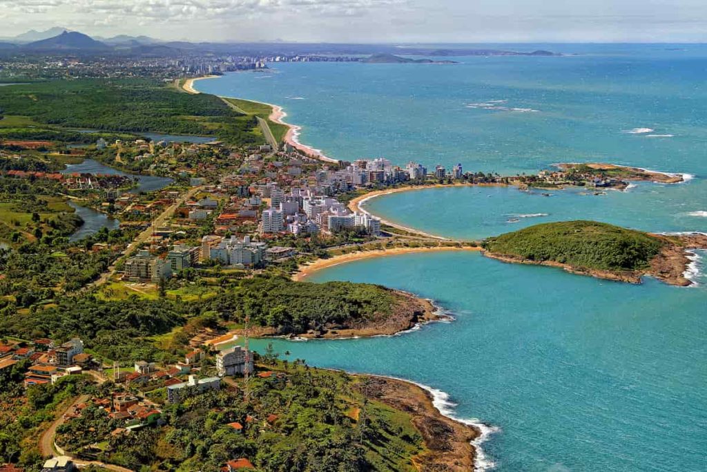 Vista aérea da Praia do Morro em Guarapari, com mar azul, faixa de areia e prédios ao fundo em dia ensolarado.