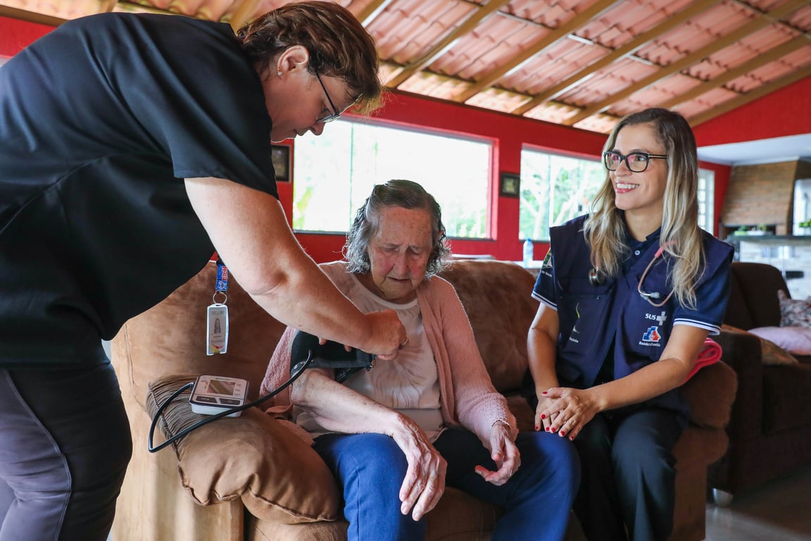 Agente comunitário de saúde em visita domiciliar, medindo pressão arterial em ambiente residencial simples