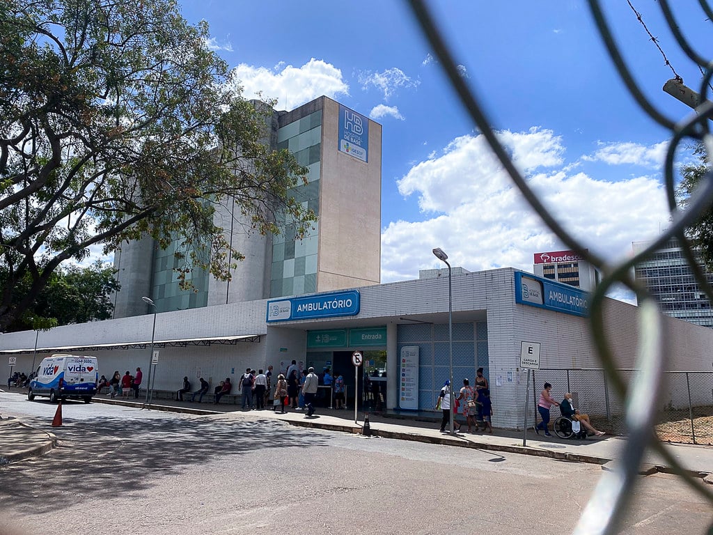 Fachada do Hospital de Base do Distrito Federal (HBDF), em Brasília, com movimento de pessoas na entrada e céu parcialmente nublado.