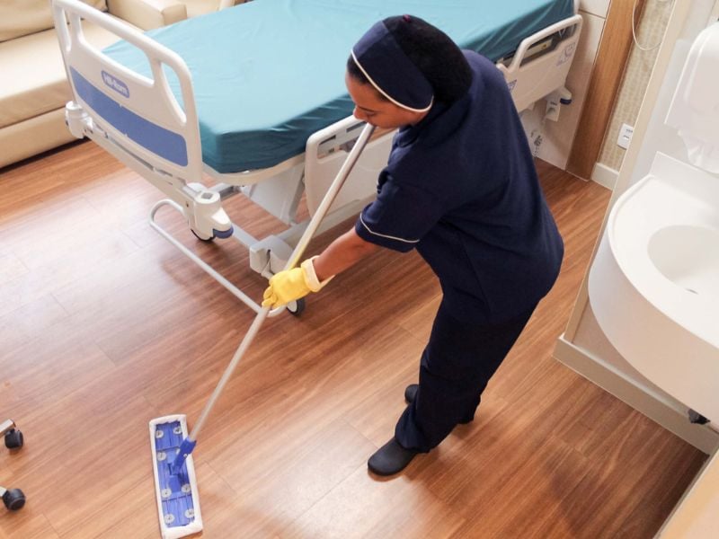 Profissional de limpeza em corredor hospitalar, próximo a leito