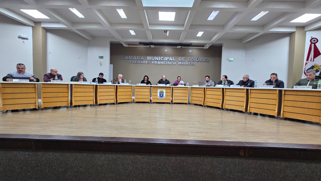 Interior de plenário de câmara municipal com mesas e cadeiras dispostas em U e brasão ao fundo, sem pessoas