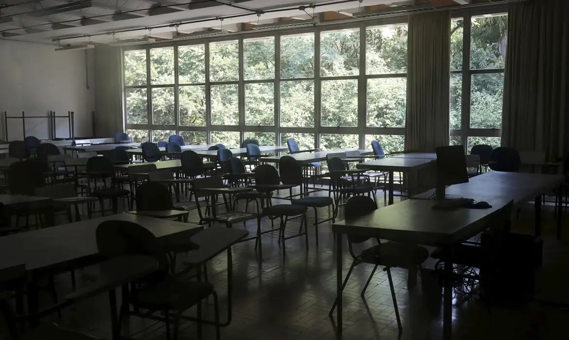 Sala de aula em escola pública, com mesas e cadeiras organizadas e iluminação natural ao fundo.