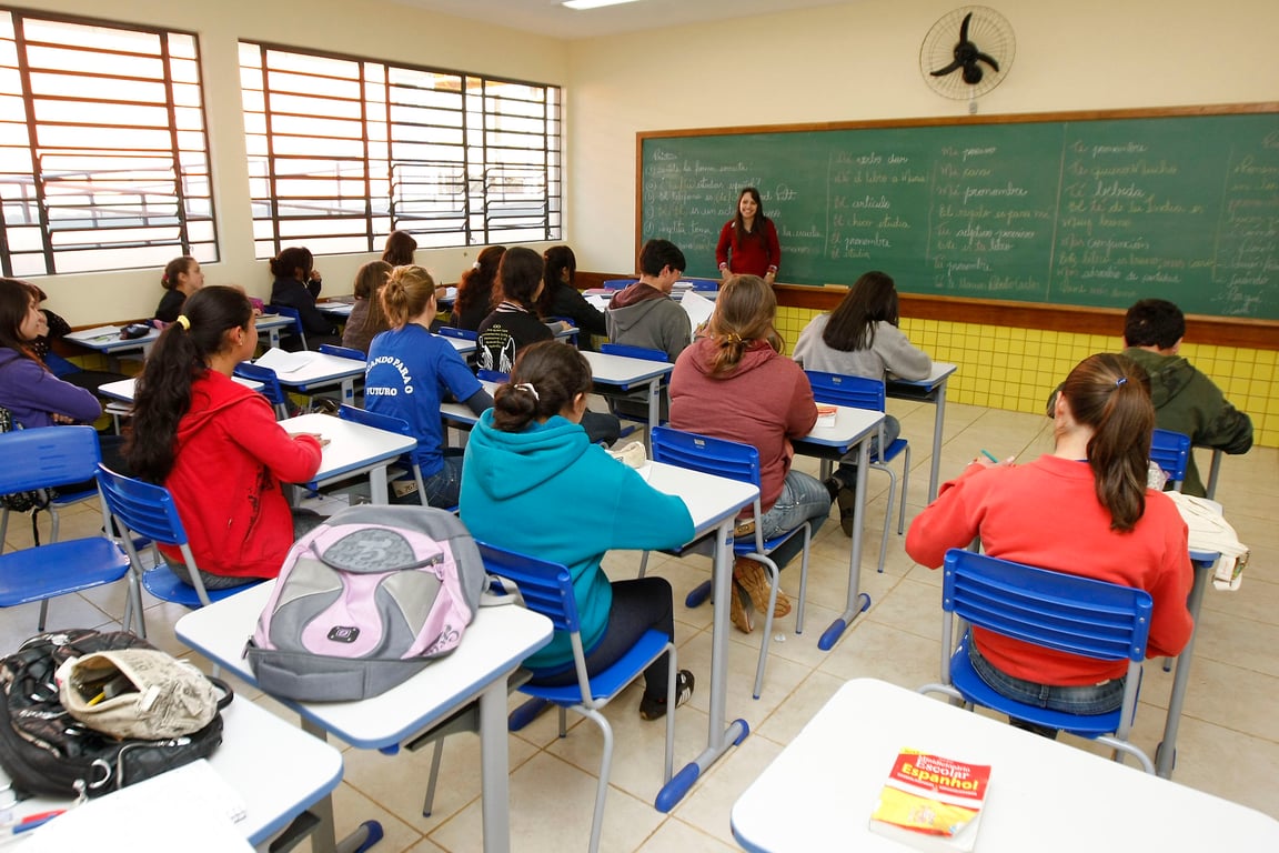 Sala de aula com estudantes e professora em frente à lousa, em ambiente escolar