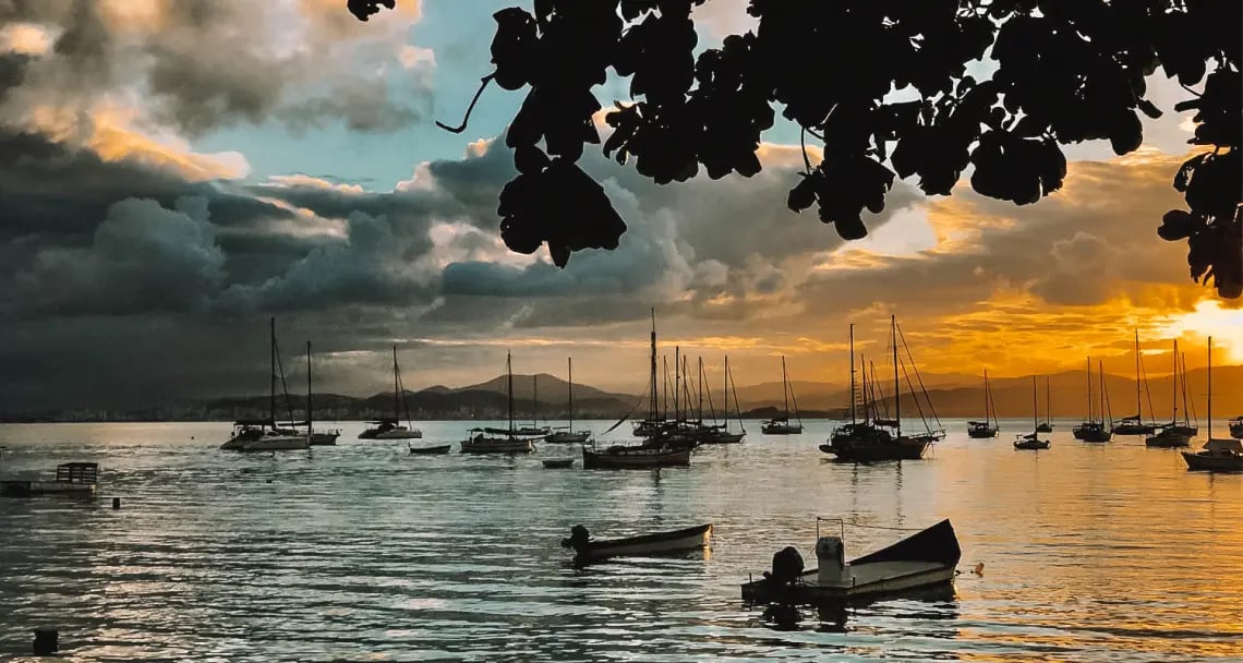Orla de Florianópolis ao pôr do sol, com barcos e silhueta da cidade ao fundo