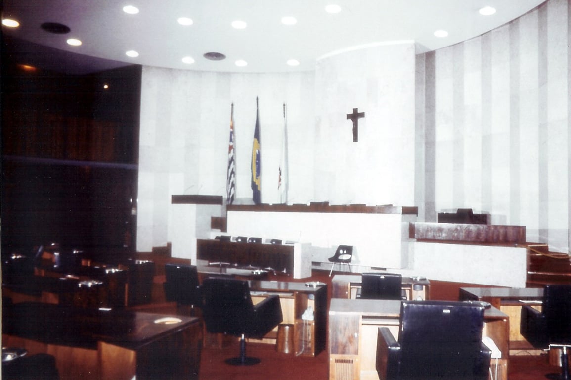 Plenário de uma câmara municipal, com cadeiras e microfones, em ambiente iluminado