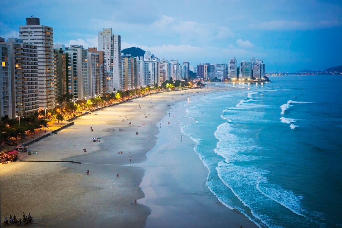 Orla do Guarujá ao entardecer, com prédios à beira-mar e faixa de areia movimentada