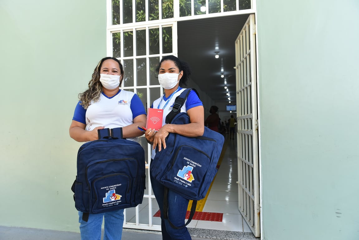 Duas agentes comunitárias de saúde com mochilas e pranchetas em ambiente de atendimento