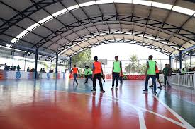 Alunos praticando corrida em aula de educação física em quadra coberta; usam coletes esportivos coloridos.