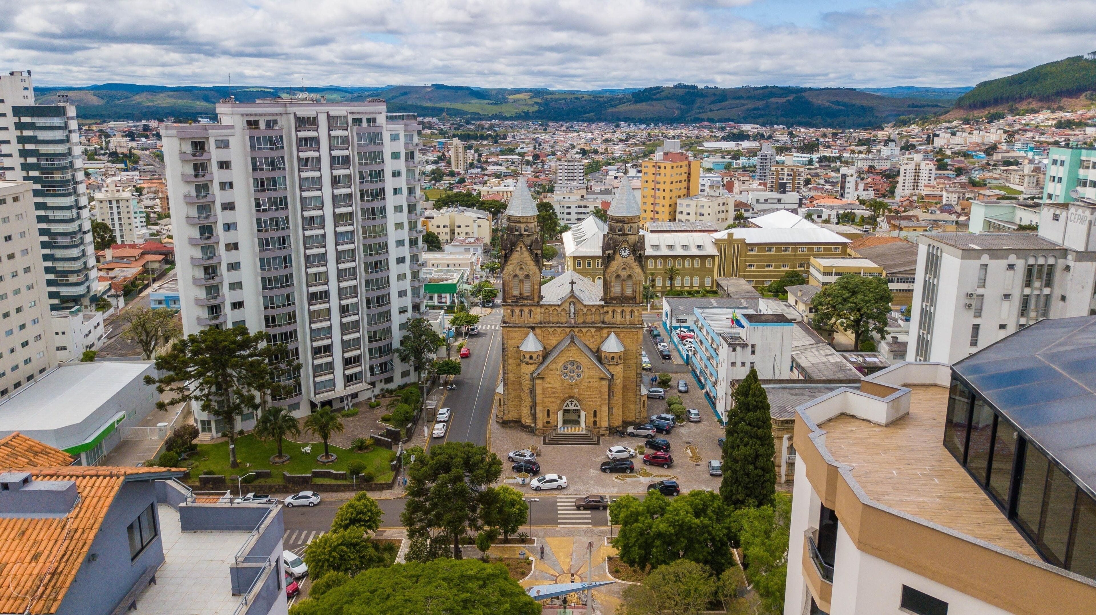 Vista aérea de cidade serrana com igreja e casario, céu azul e entorno verde, referência ao ambiente urbano da Serra Catarinense