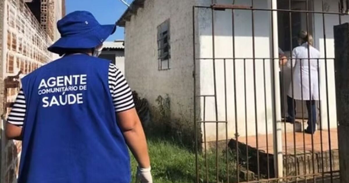 Agente comunitário de saúde caminhando em área residencial simples, com colete azul, em cenário que remete à atenção primária do SUS