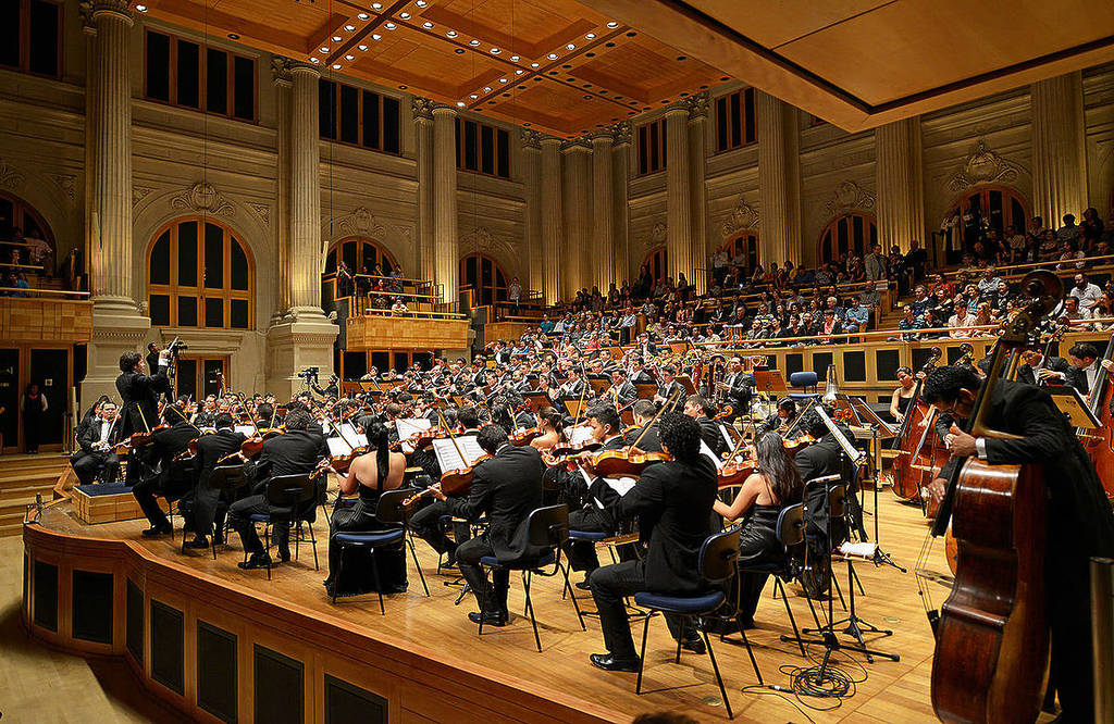 Cena de ensaio de orquestra sinfônica em auditório de São Paulo, com maestro e naipe de cordas em destaque