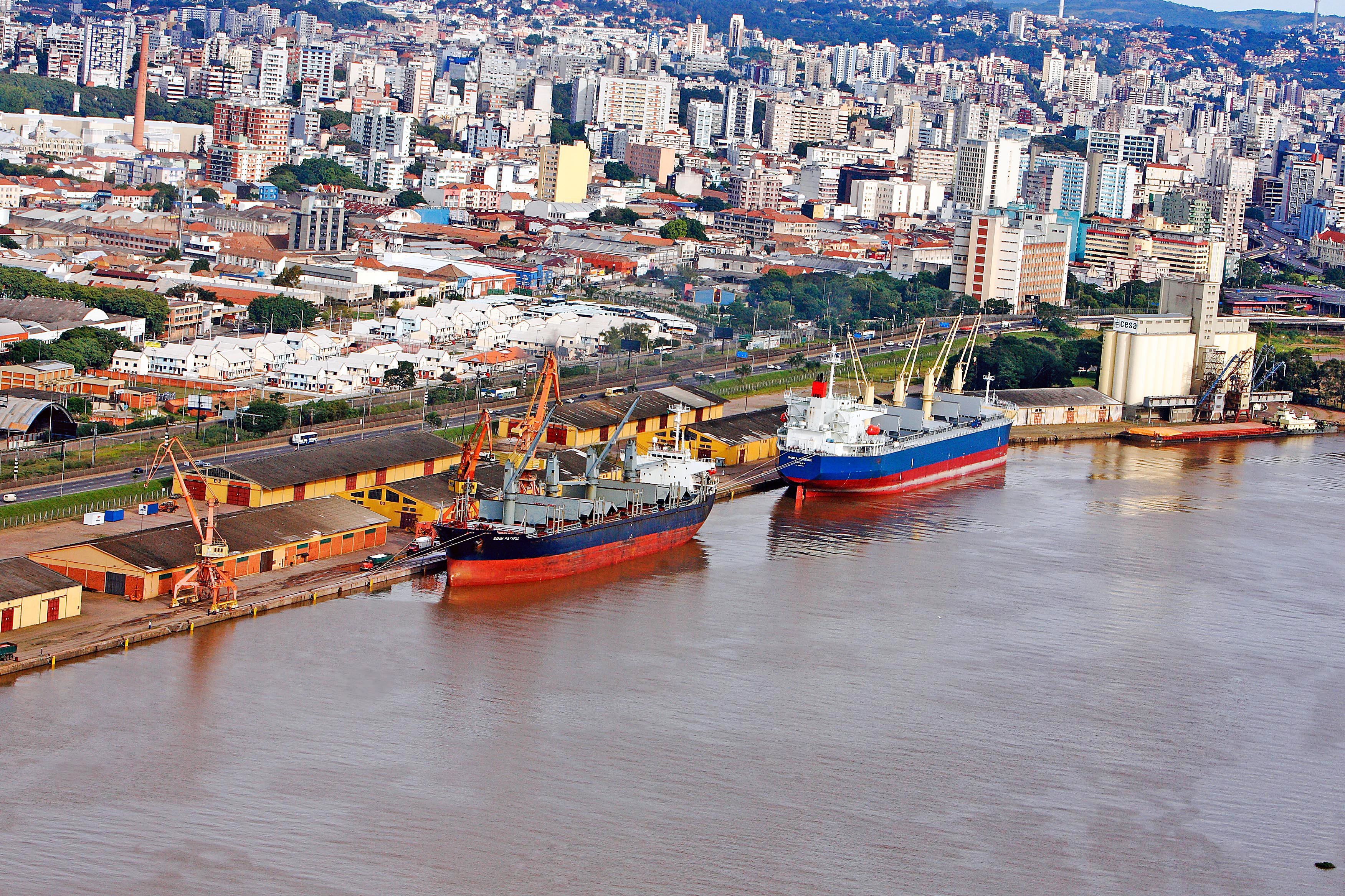Vista aérea do Porto do Rio Grande, com navios atracados e a cidade ao fundo, em dia ensolarado.
