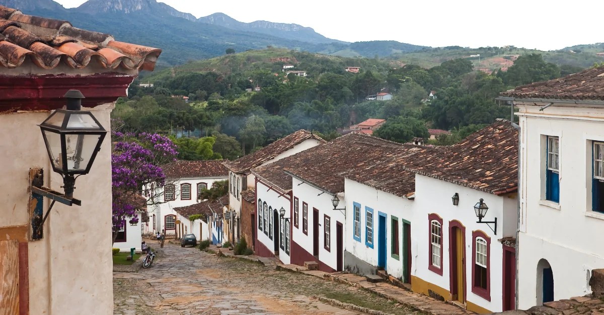 Rua típica de cidade do interior de Minas Gerais, com casario baixo e montanhas ao fundo, em fim de tarde