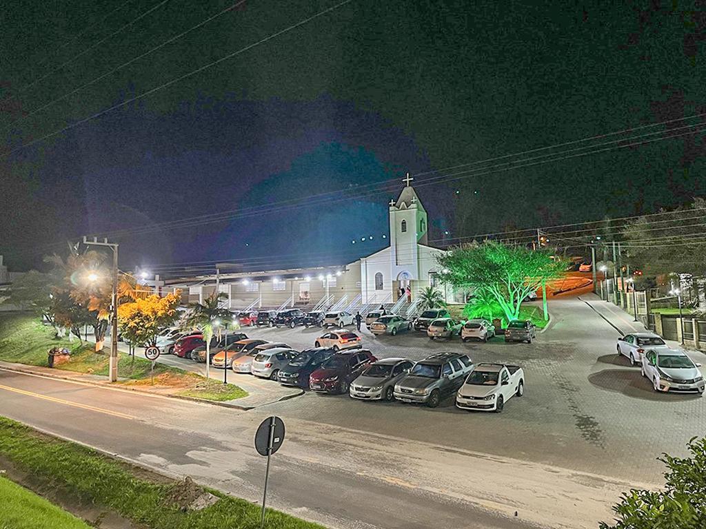 Centro urbano de cidade do Sul catarinense, com igreja iluminada ao fundo e movimento de veículos à noite