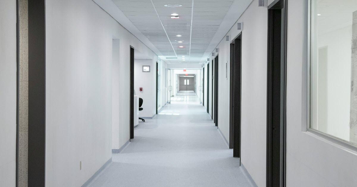 Corredor hospitalar com portas, sinalização e iluminação, representando a logística e o apoio administrativo