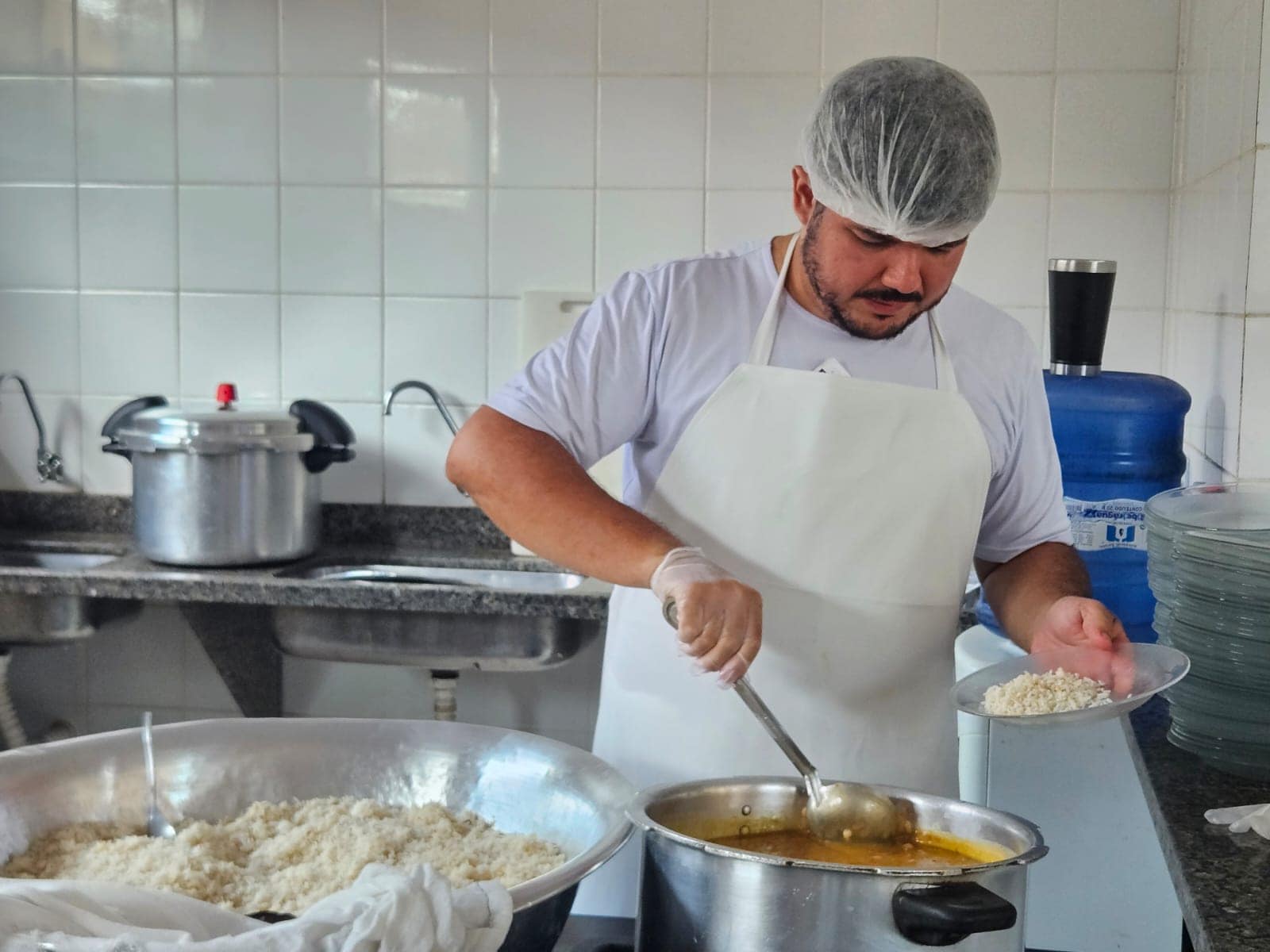 Cozinha industrial com profissional usando EPIs (touca e luvas) preparando alimentos