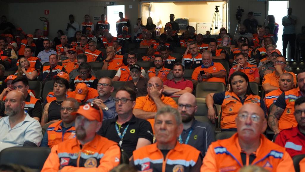 Equipe da Defesa Civil em capacitação, com uniformes laranja, sentada em auditório durante treinamento