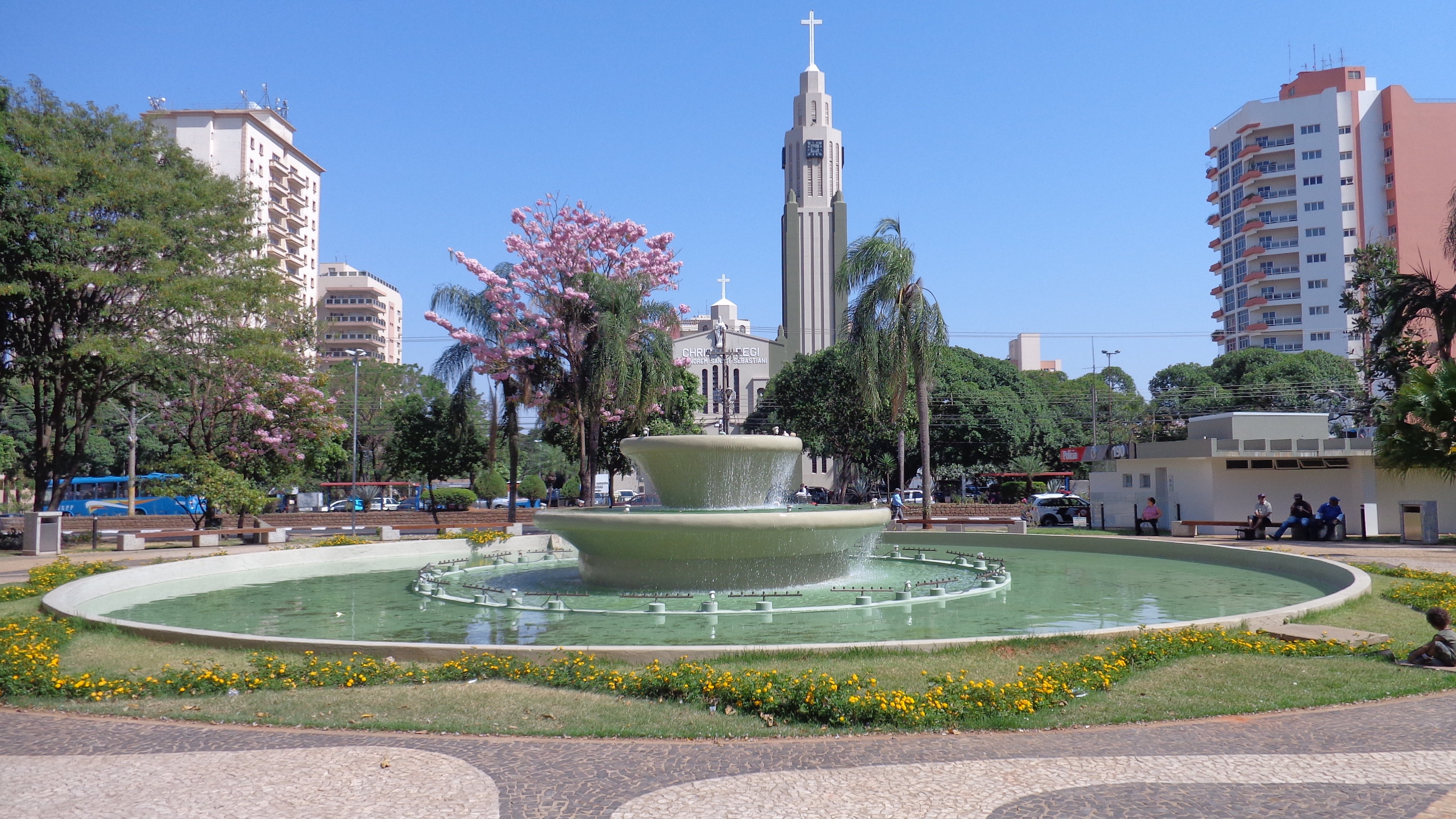 Praça arborizada de cidade do interior paulista, com igreja ao fundo
