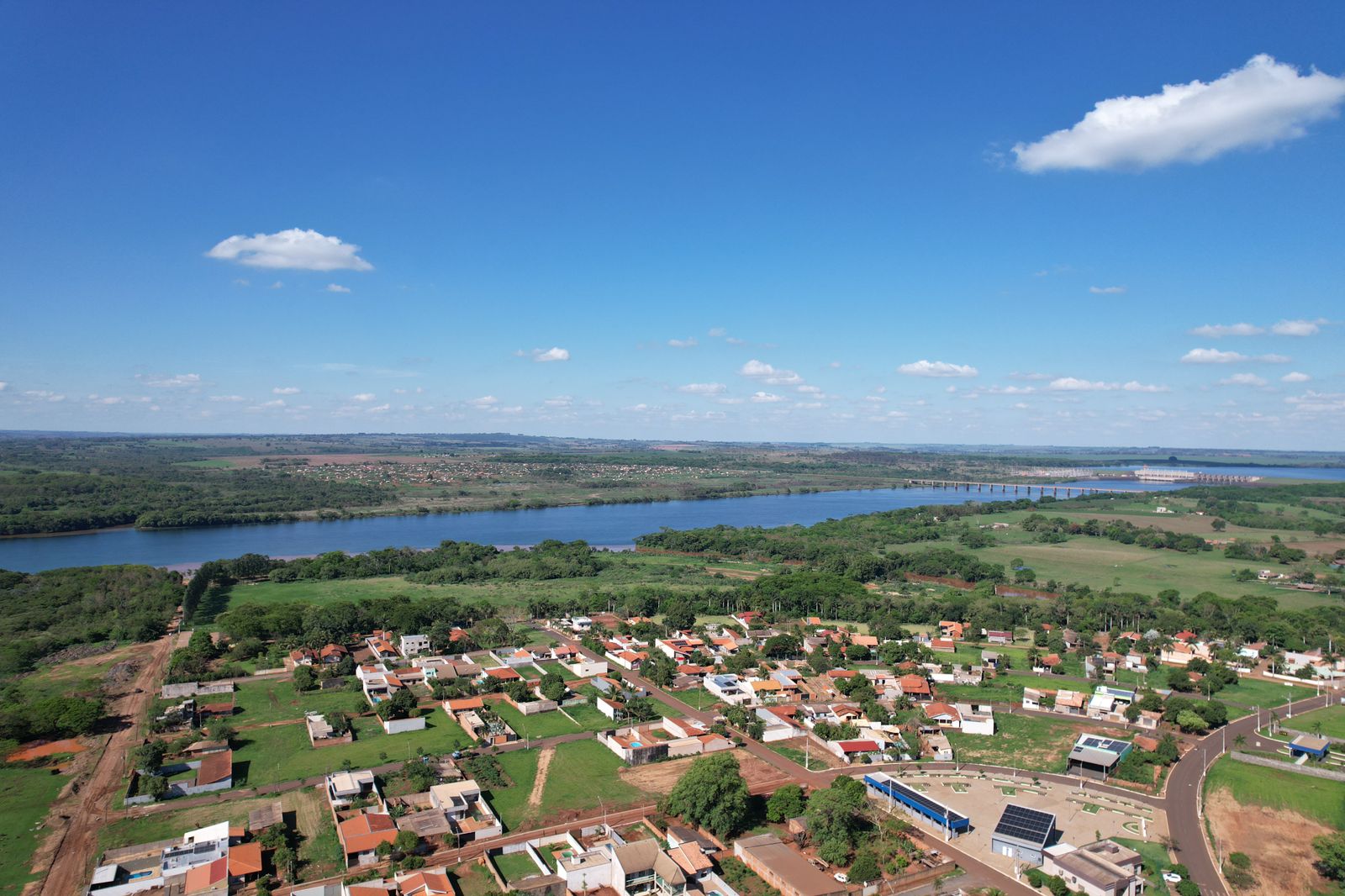 Vista aérea de cidade pequena com áreas verdes e rio ao fundo, céu aberto e nuvens brancas